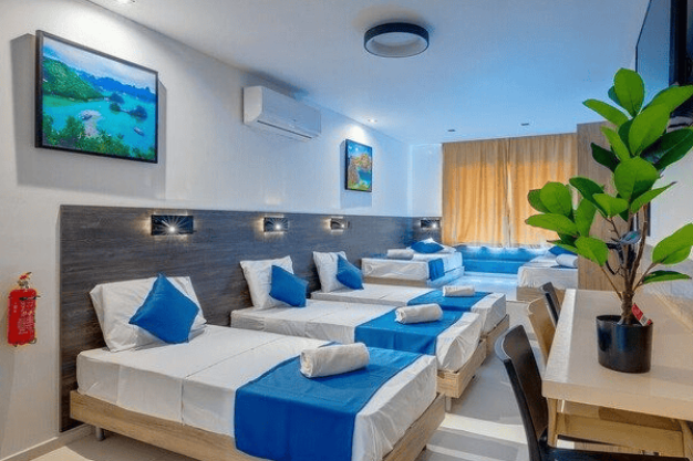 Spacious hotel room with multiple single beds, blue bedding accents, work desk, air conditioning and modern amenities for comfortable group accommodation.