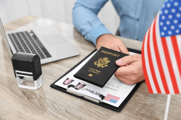 Person holding a United Kingdom passport over a U.K. visa application form with American flag, laptop, and passport stamp during visa processing.