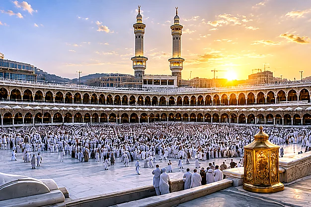 Sunset view of the Grand Mosque in Makkah with two minarets and pilgrims gathered in the courtyard.