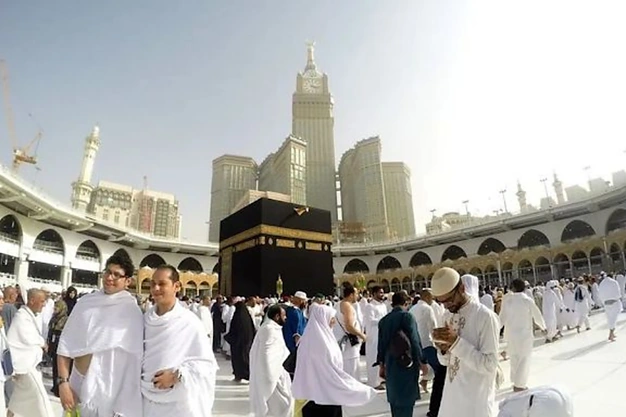 Ground-level view of the Kaaba at Masjid al-Haram in Makkah with pilgrims in Ihram performing Umrah and the Abraj Al Bait Clock Tower in the background.