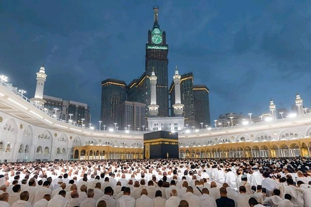 Nighttime view of the Kaaba at Masjid-al-Haram with worshippers in prayer and Abraj Al Bait Clock Tower and mosque minarets rising with lights