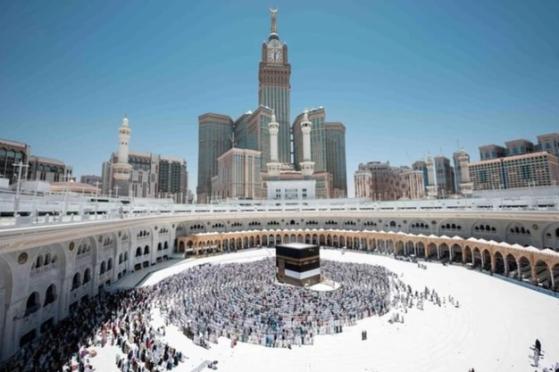 Wide view of the Kaaba at the center of Haram surrounded by worshippers with the iconic Abraj Al Bait Clock Tower rising prominently against a clear blue sky.