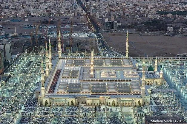 Aerial view of Al-Masjid e Nabawi in Madinah featuring the Green Dome, illuminated courtyards and thousands of worshippers gathered for prayers.
