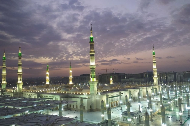Evening view of Al-Masjid-an-Nabawi in Madinah with minarets, courtyards and a dramatic twilight sky.