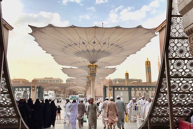 Courtyard of Al-Masjid-e-Nabawi in Madinah with iconic retractable umbrellas and Muslim pilgrims visiting during their Umrah journey.
