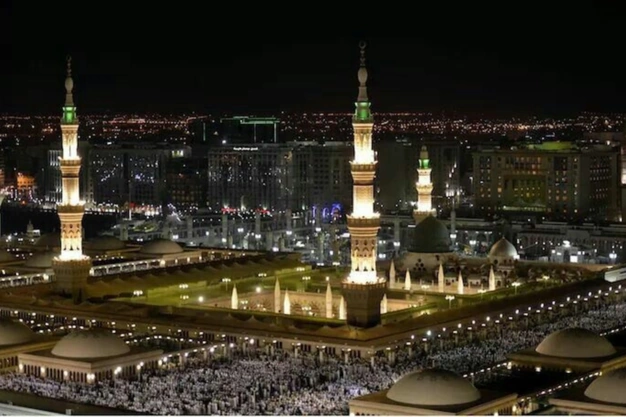 Night view of Al-Masjid Nabawi featuring the Green Dome, beautiful minarets and worshippers gathered in the mosque courtyards.