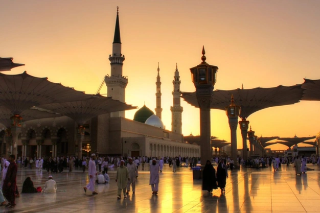 Golden sunset view of Masjid Nabawi with iconic Green Dome between minarets, worshippers and open umbrellas silhouetted against the evening sky.
