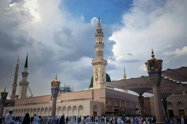 Pilgrims at Masjid-e-Nabawi in Madinah with the iconic Green Dome, towering minaret, and mosque courtyard beneath a dramatic cloudy sky.