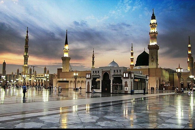 Al Masjid Nabawi in Madinah at sunset featuring the Green Dome, minarets and pilgrims in the mosque courtyard.