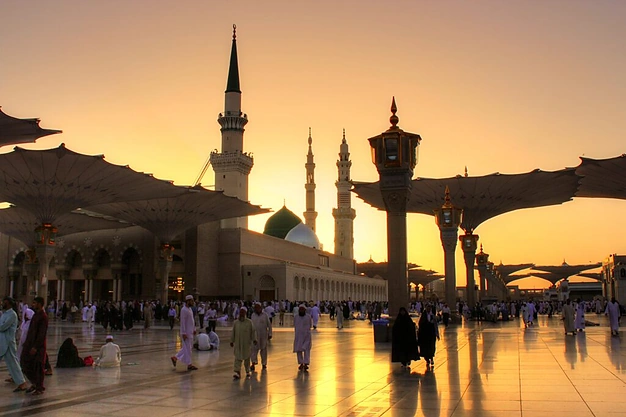 Golden sunset view of Masjid-e-Nabawi with the Green Dome, open courtyard umbrellas, and worshippers in Madinah.