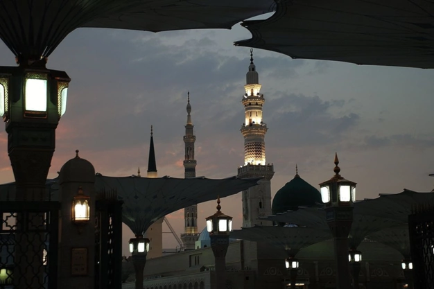 Dusk view of Masjid e Nabawi in Madinah, with minarets, the iconic green dome, and large open courtyard umbrellas beneath a twilight sky.