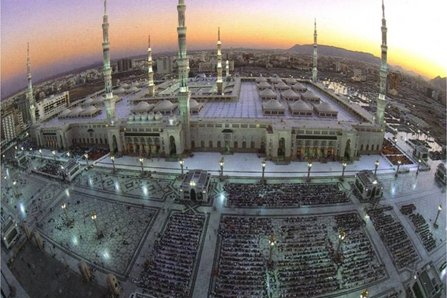 Aerial sunset view of Al-Masjid e Nabawi in Madinah with worshippers gathered in the mosque courtyard and iconic illuminated minarets.