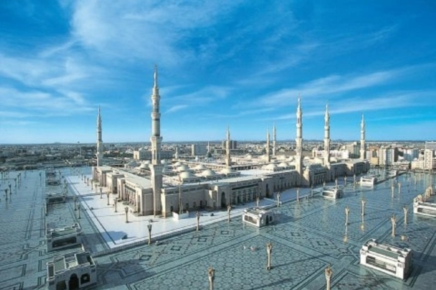 Panoramic aerial view of The Prophet's Mosque featuring iconic minarets, courtyard and surrounding cityscape under a clear blue sky.